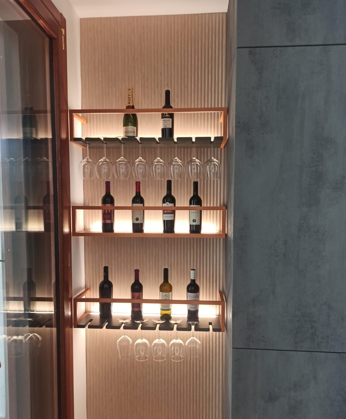 Portabottiglie incassato nel muro, con bottiglie e bicchieri. Ripiani in legno retroilluminati su una parete strutturata.