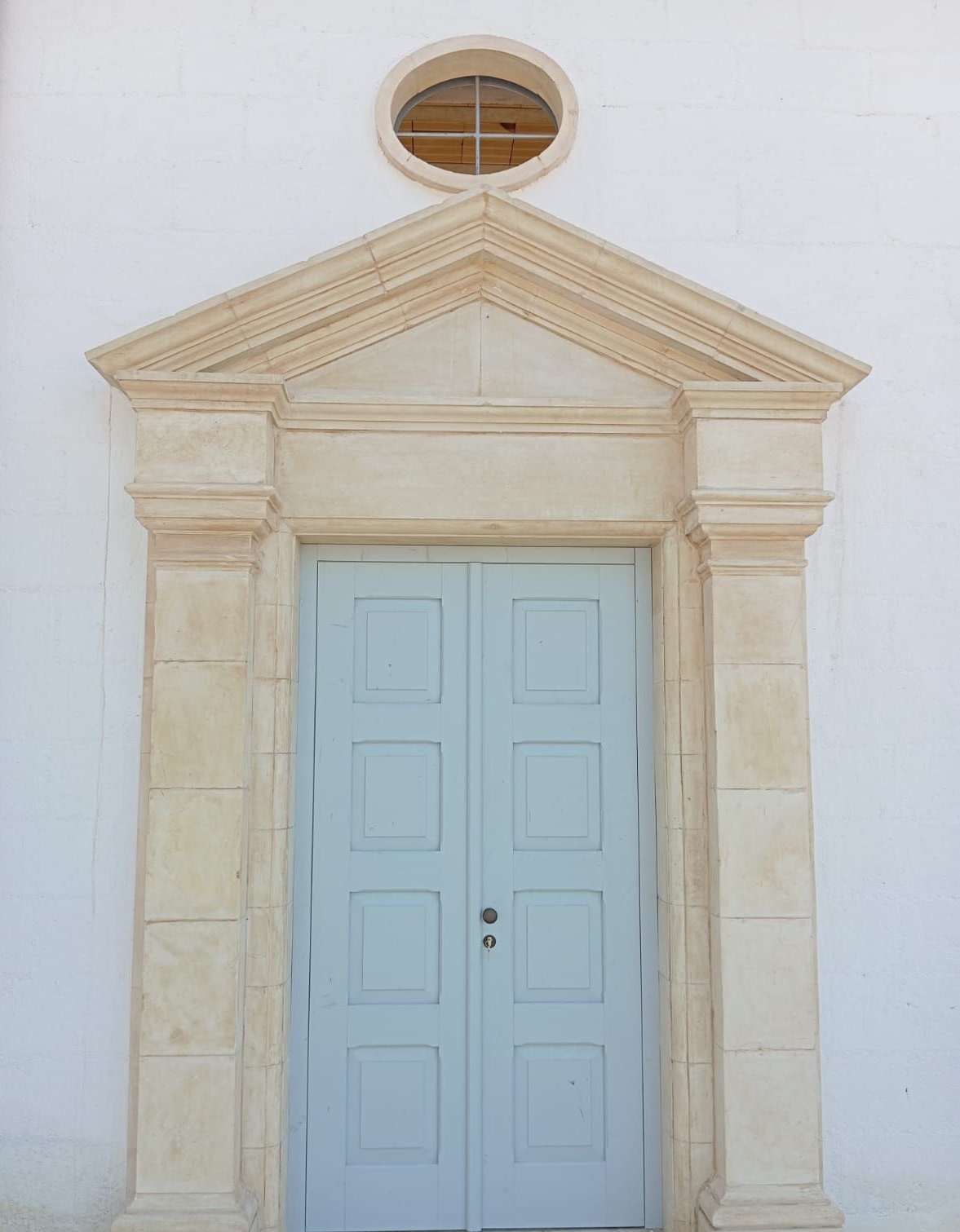 Doppie porte azzurre sotto una cornice in pietra chiara e un frontone triangolare, con una finestra ovale sopra.