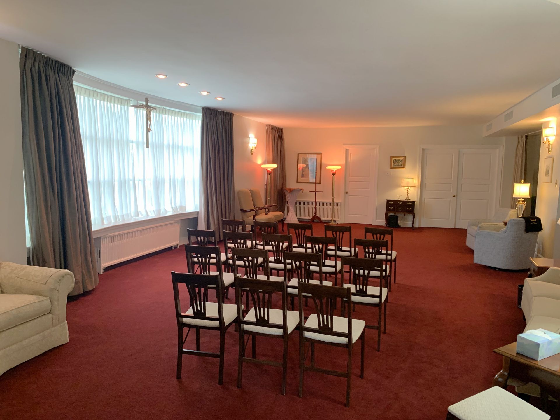 Interior of a funeral home chapel with chairs arranged for a service, red carpet, and a cross on the wall.