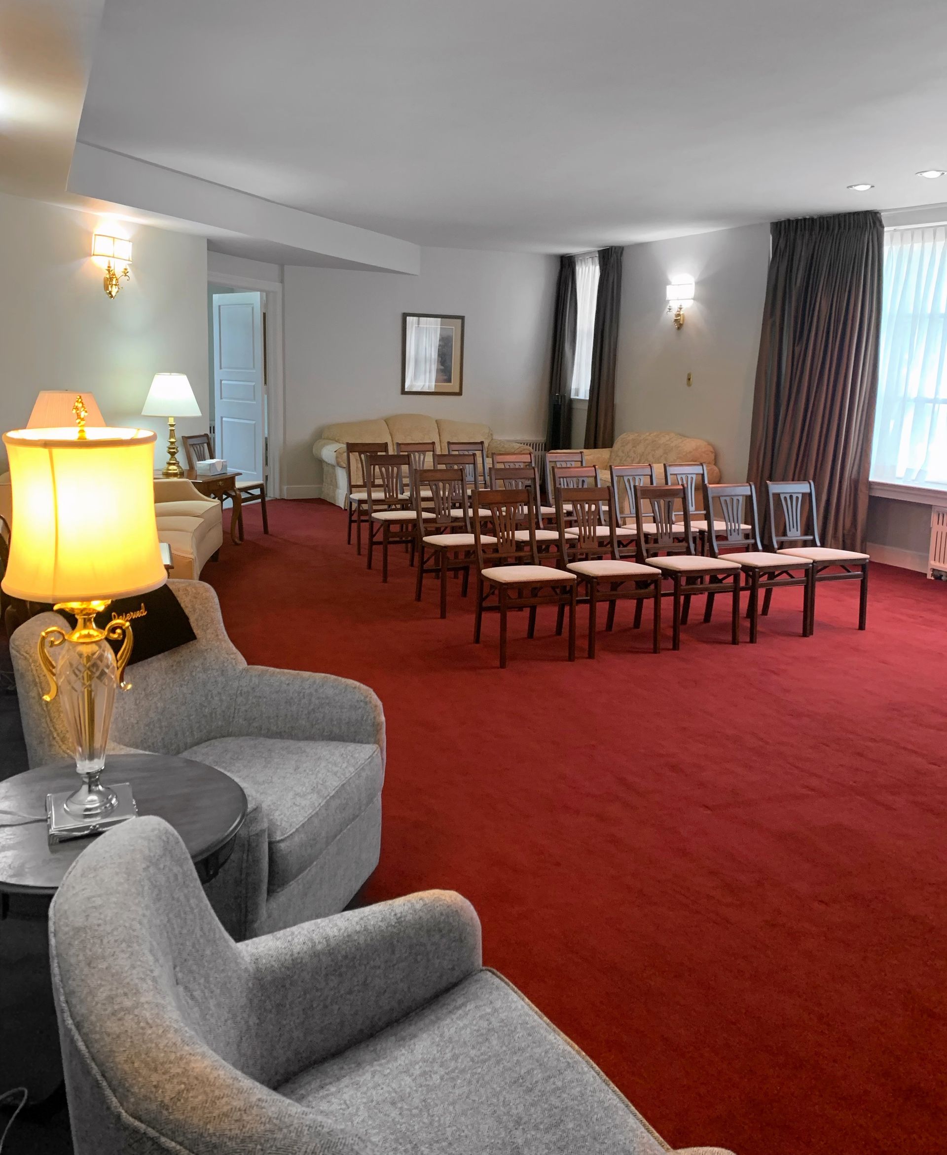 An empty room with a red carpet, chairs arranged in rows, and armchairs near a lit lamp.