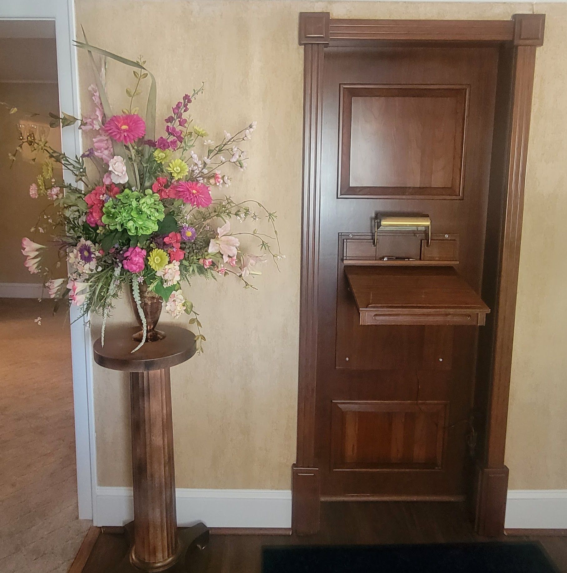 Flower arrangement beside a dark wooden phone booth-style cabinet in a room with light yellow walls.