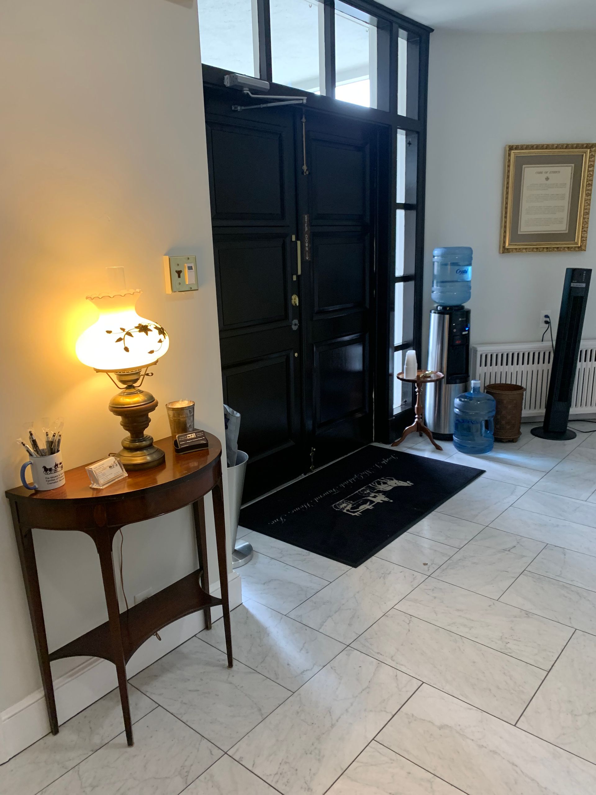 An entryway with a dark door, marble floor, a table with a lamp, and water dispenser.