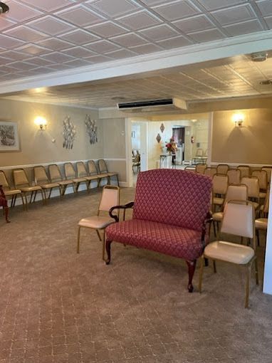 Interior room with rows of chairs and a patterned loveseat; beige walls, carpet, and ceiling tiles.