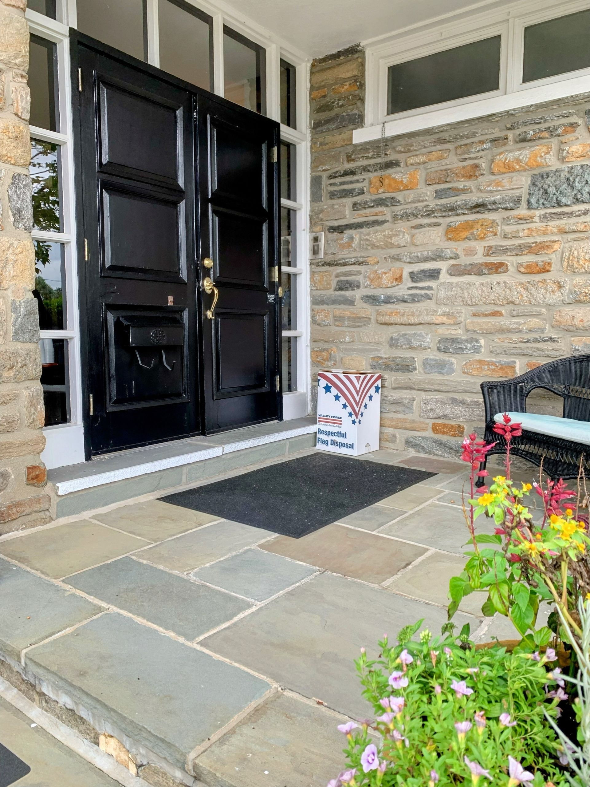Black double doors on a stone porch. A mat is in front of the doors, with a package and flowers nearby.