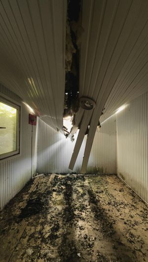 An interior room with white, paneled walls and a collapsing ceiling, exposing debris on the dark floor below.
