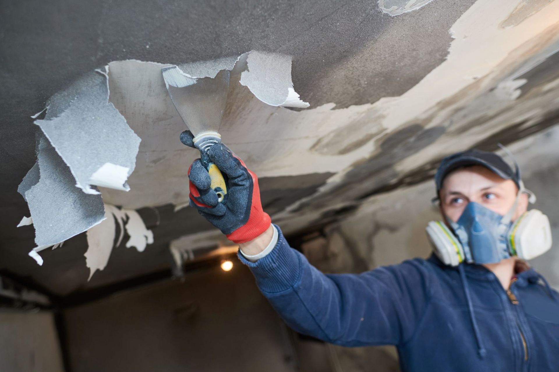 A person wearing a respirator mask uses a putty knife to scrape peeling paint off a ceiling.