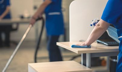 A cleaning professional in a blue uniform wipes a table, while another vacuums in the background of an office.