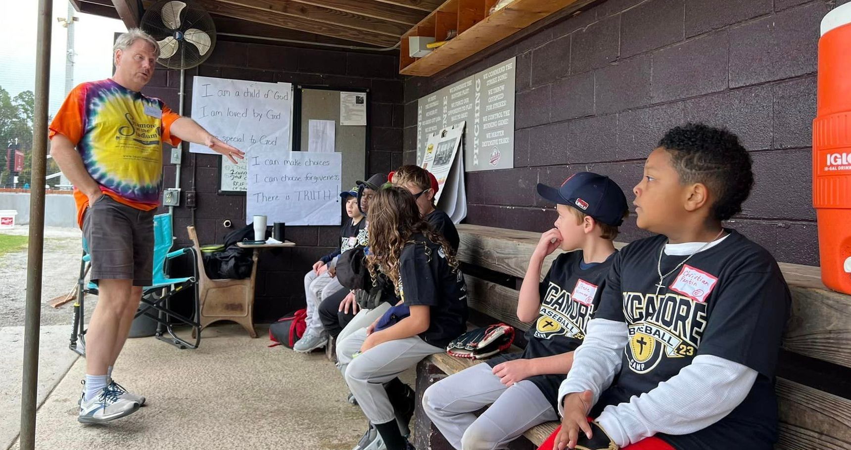 A man in tie-dye shirt shares points with seated children wearing baseball t-shirts.