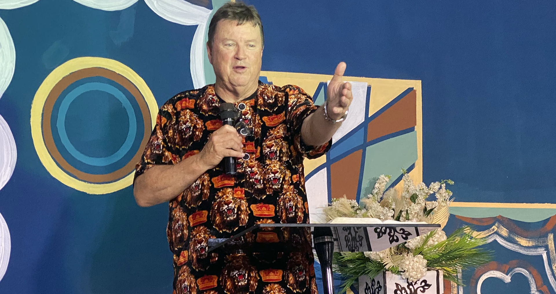 Man speaking at a podium, wearing a colorful patterned shirt, gesturing with his right hand; blue backdrop.