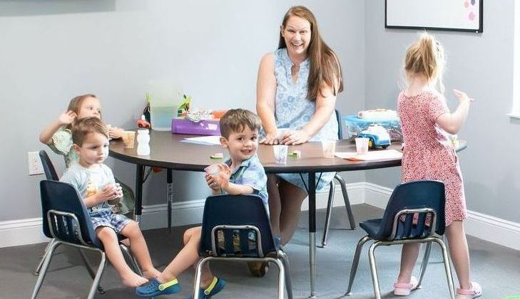 A teacher smiles at children seated around a table in a classroom.