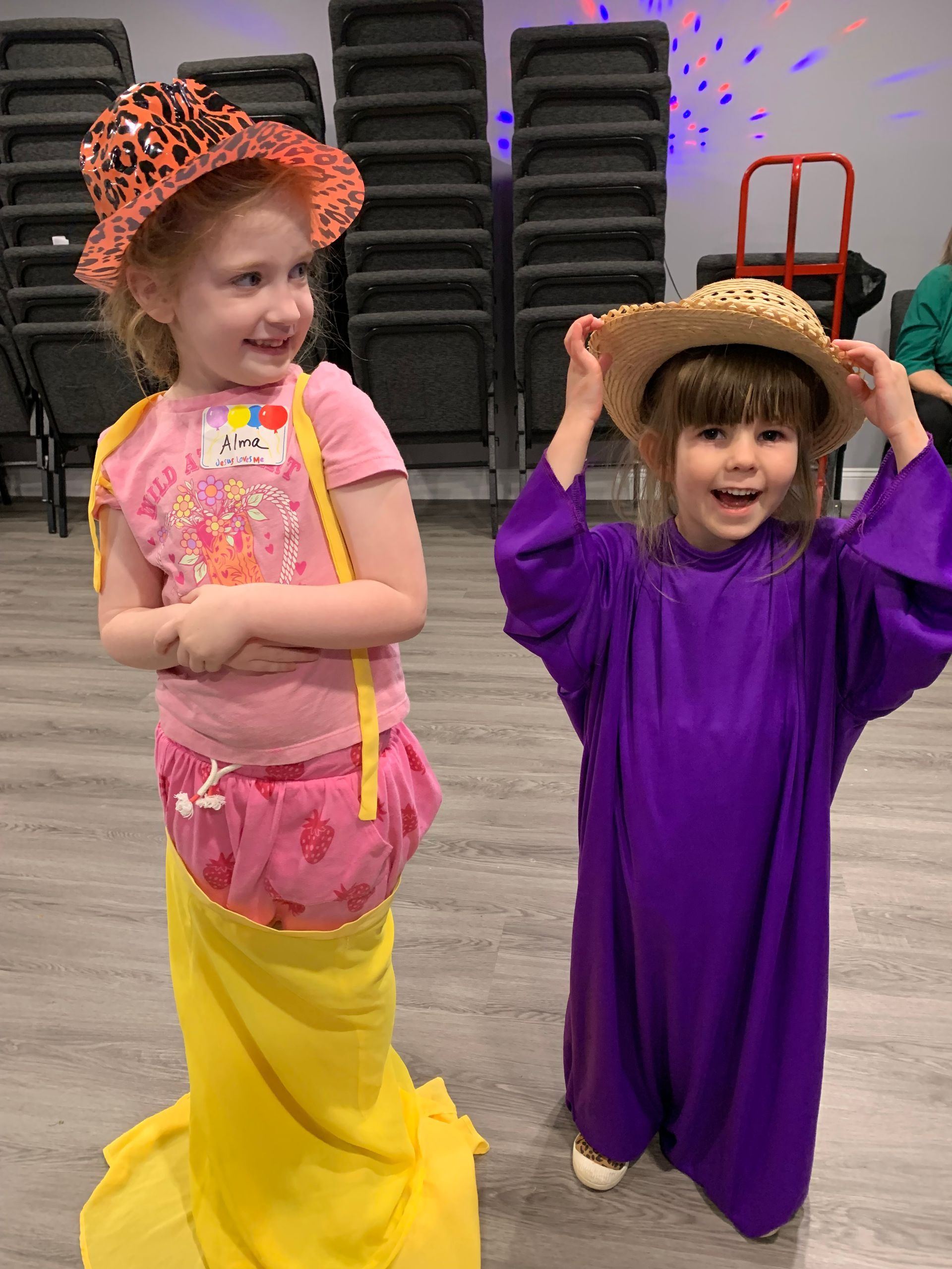 Two young girls in costumes, one in pink and yellow, the other in purple, wearing hats, smiling.