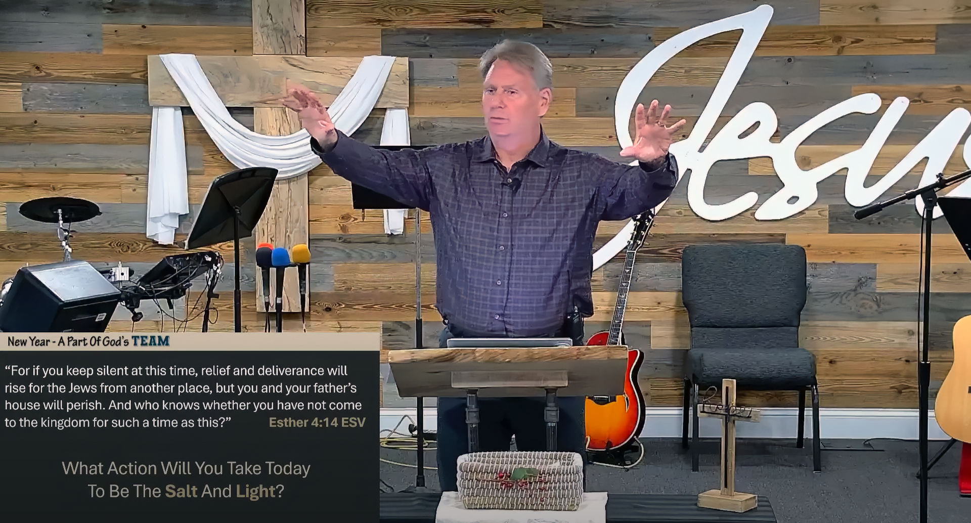 Man in a church setting gesturing, with Bible text on screen. Wooden background with guitars, chairs.