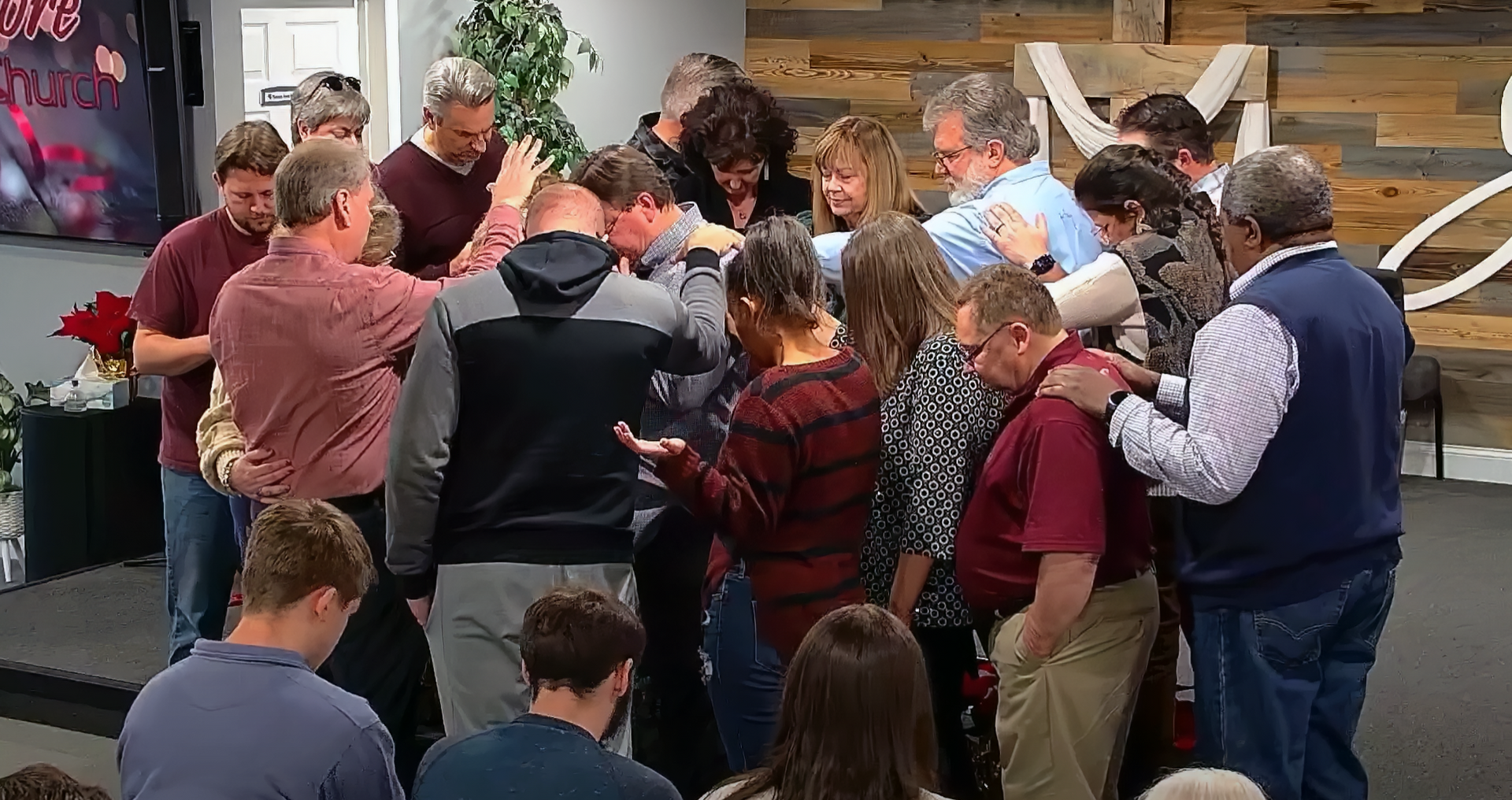 Group of people in prayer, hands on shoulders, in a church setting.