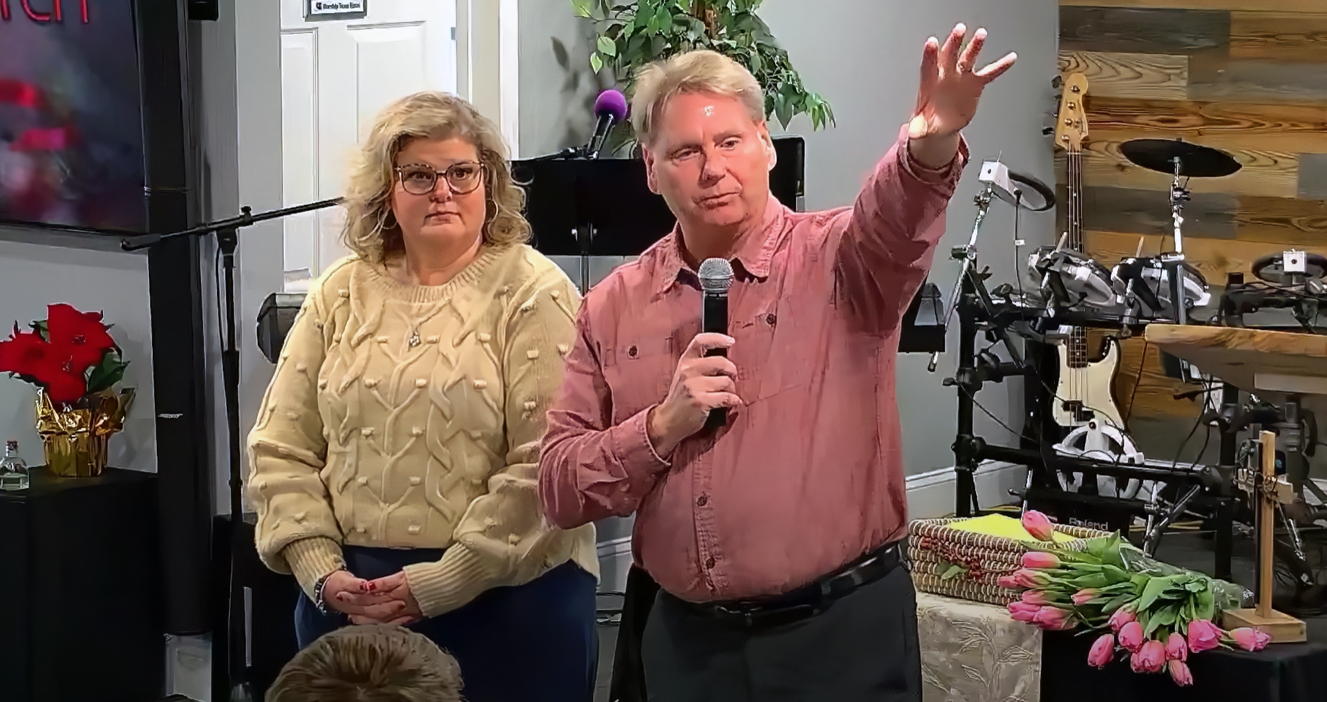Man speaking at microphone, woman beside him. Indoor stage with flowers and music equipment.