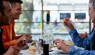 Men laughing at a table in a restaurant, with food and drinks in front of them.