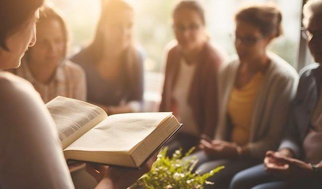 Woman reading from an open book to a group of women in a bright room, soft focus.