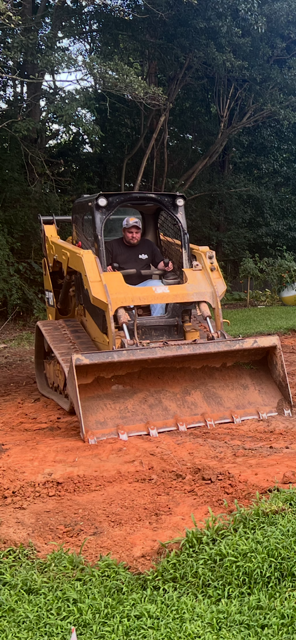 A person operating a yellow bulldozer on red dirt; green grass in the foreground. Trees in the background.