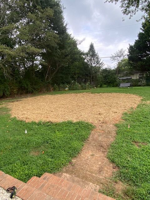 A grassy backyard with a large mulched area, surrounded by trees and a brick patio in the foreground.