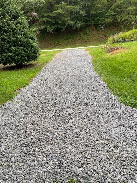 Gravel path leading toward a wooded area. Green grass borders the gray path.