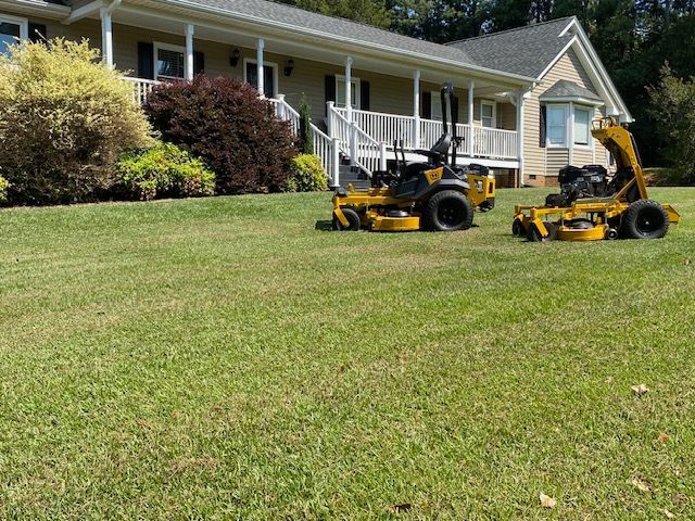 Two yellow lawn mowers parked on a green lawn in front of a house with a white porch.