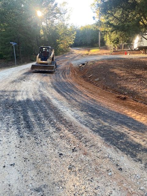 Skid steer compacting gravel road on a sunny day near trees.