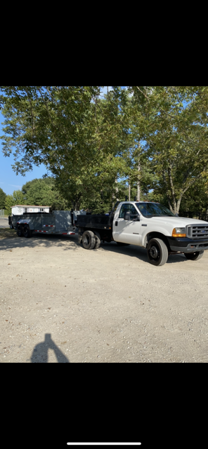 White pickup truck towing a trailer on a dirt road, under green trees, sunny day.