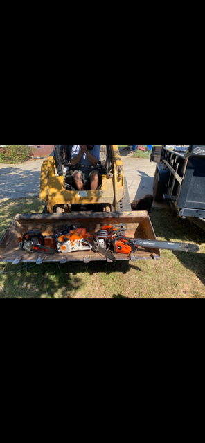 A yellow excavator carrying multiple chainsaws in its bucket.