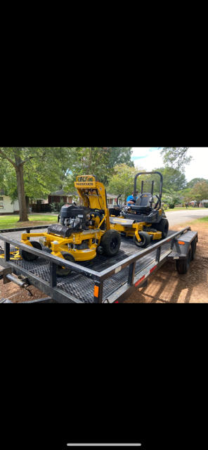 Two yellow lawn mowers on a black trailer, parked outside.