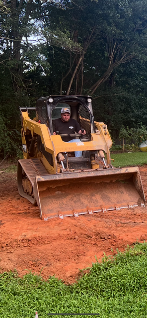 Person operating a yellow track loader on red dirt, with greenery in the foreground and trees in the background.