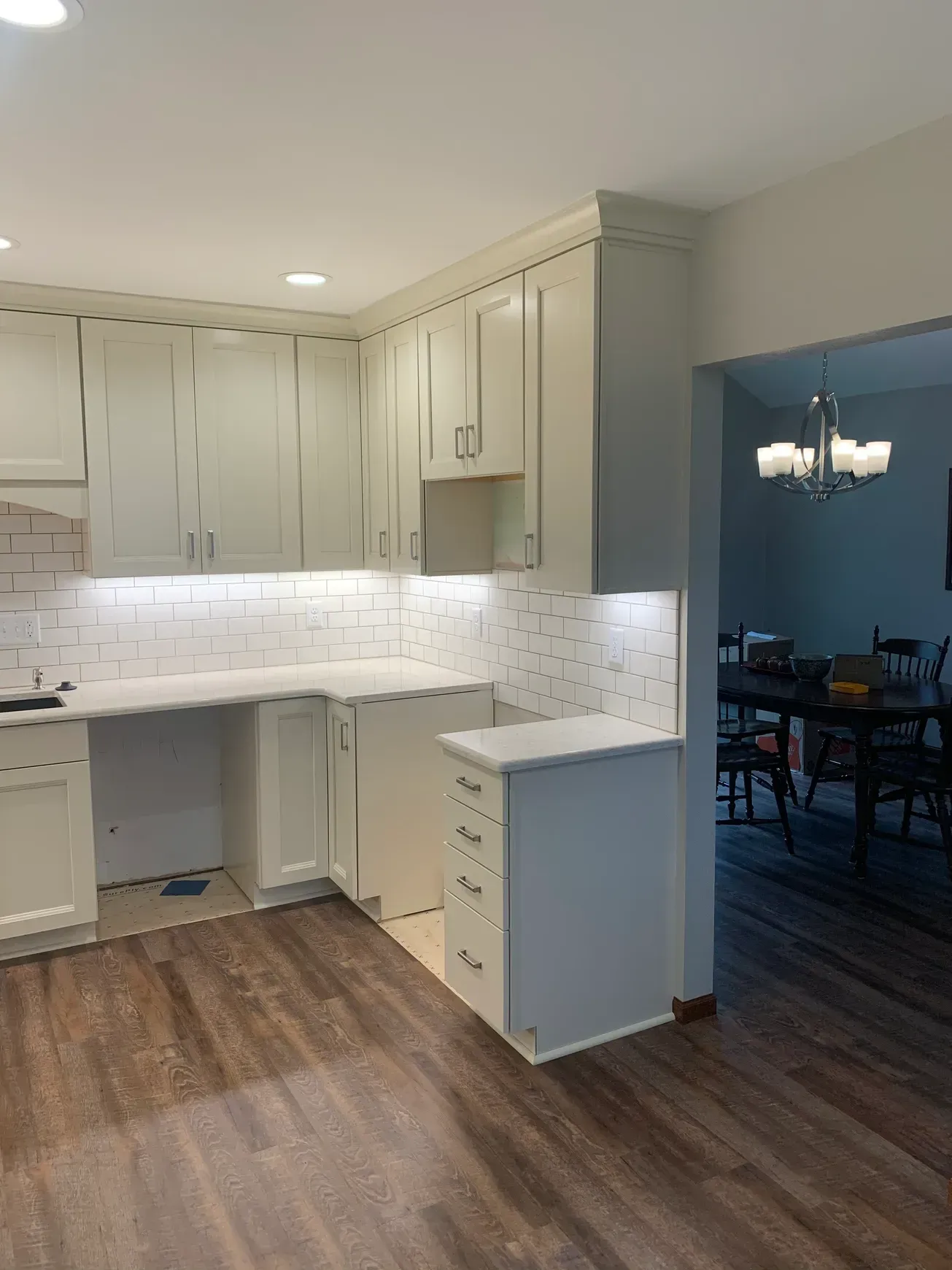 A kitchen with white cabinets and wooden floors and a dining room in the background.