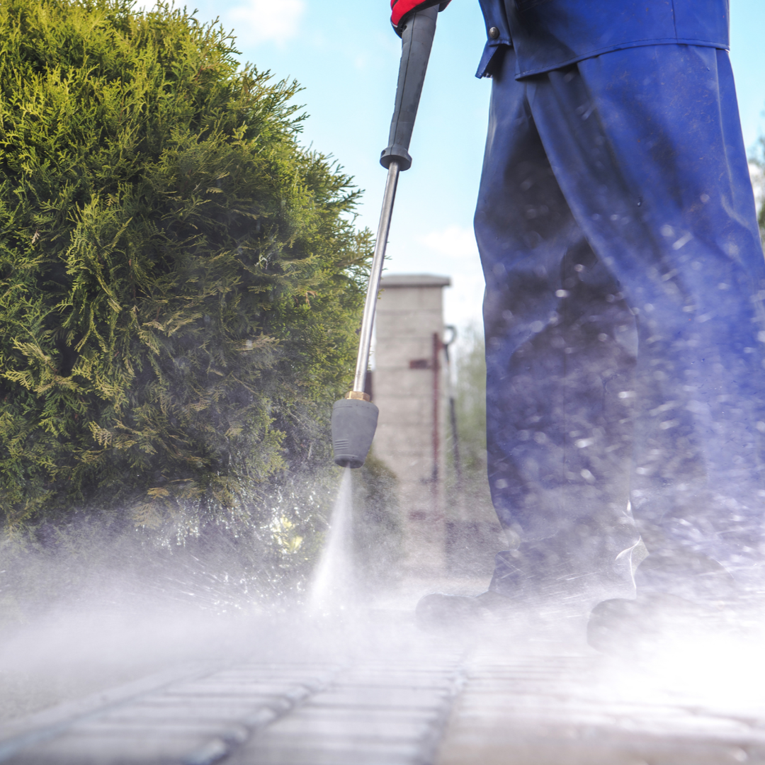 A person in blue work clothing pressure washes a stone walkway next to a leafy green bush.