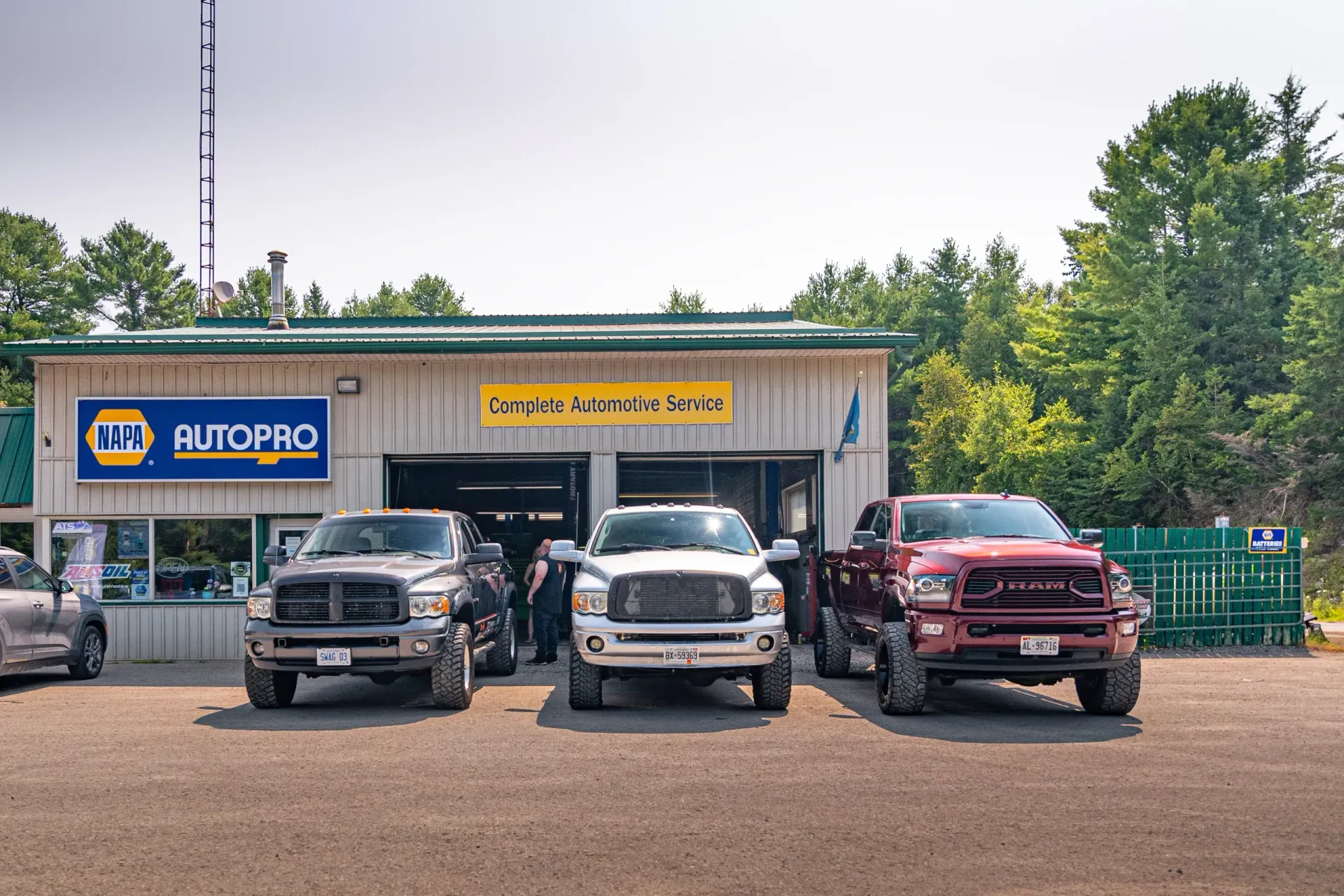 Three pickup trucks parked in front of a NAPA Auto Pro shop with a gravel lot and surrounding trees.