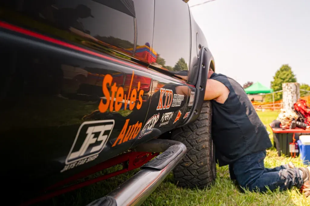 A person kneels in the grass to work on the tire of a black truck with branded decals parked in an outdoor field.