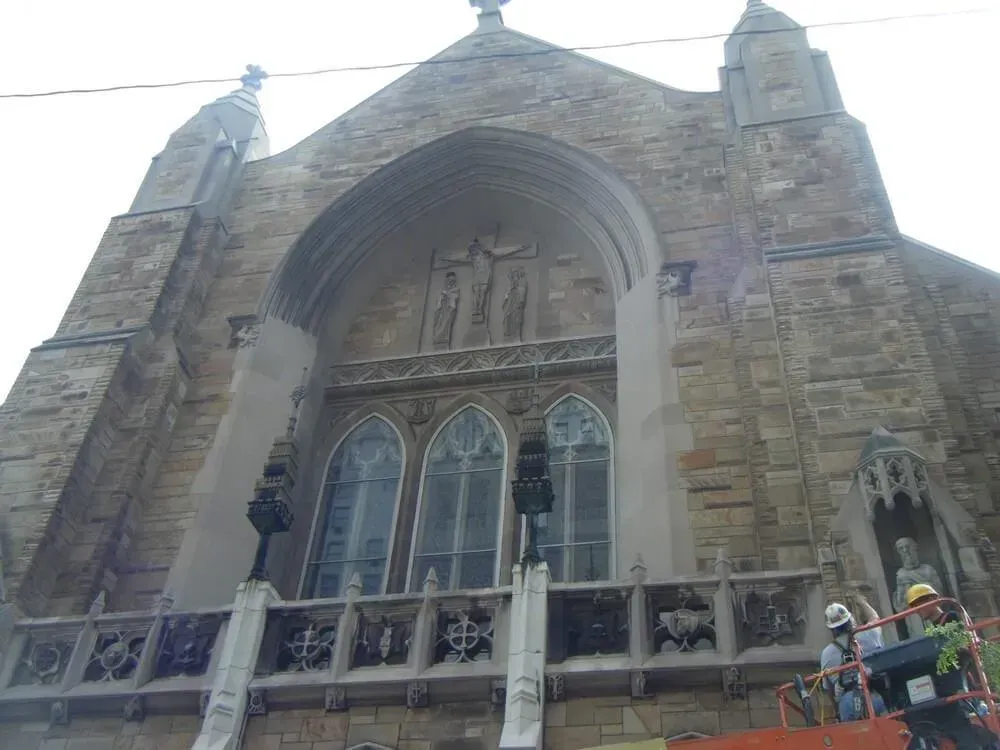 Stone church exterior with arched entrance, stained glass windows, and construction workers on lift.