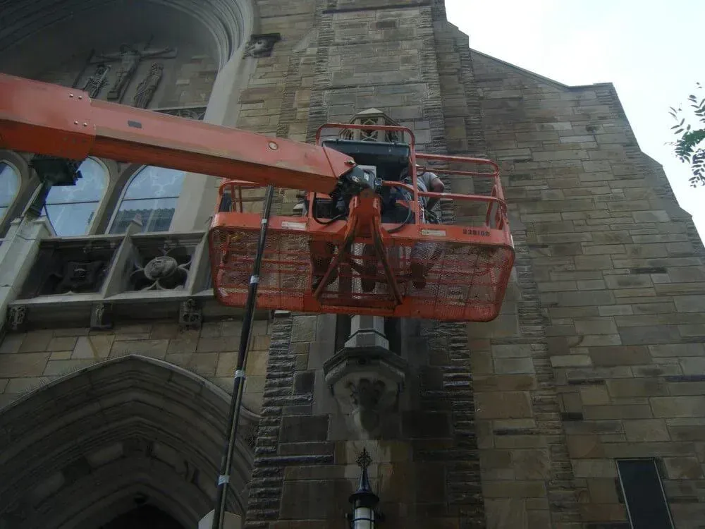 Workers in an orange lift platform repairing stonework on a tall, aged building.