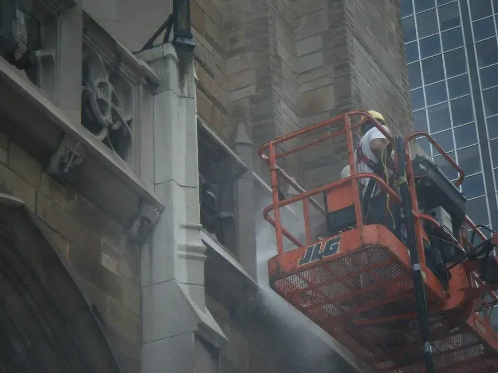 Worker on lift washing a tall stone building with a high-rise in the background.