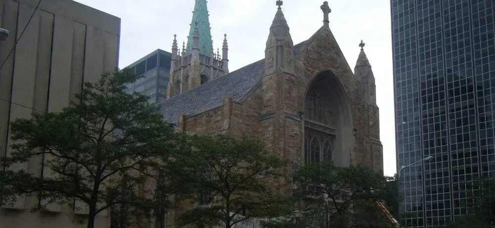 Gothic-style church with a green spire, trees, and modern buildings in an urban setting.