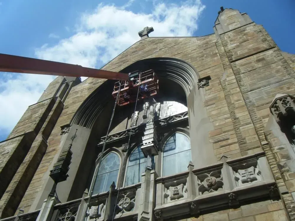 Workers in lift repairing arched window of a large stone church. Blue sky overhead.