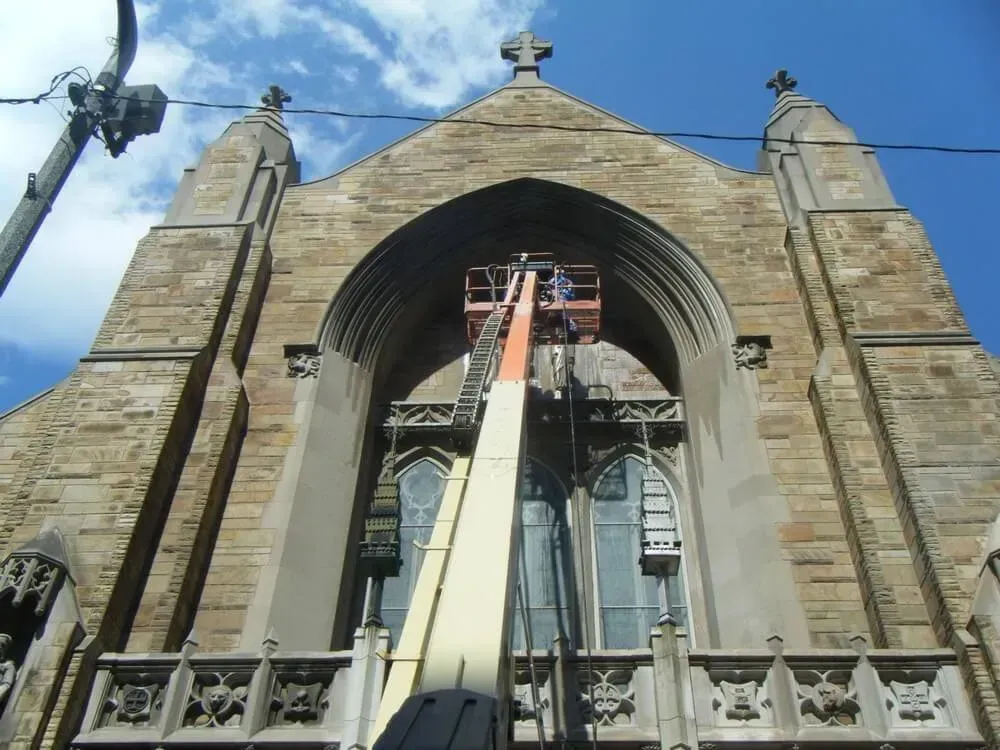 A person in a lift working on the facade of a stone church with arched windows.