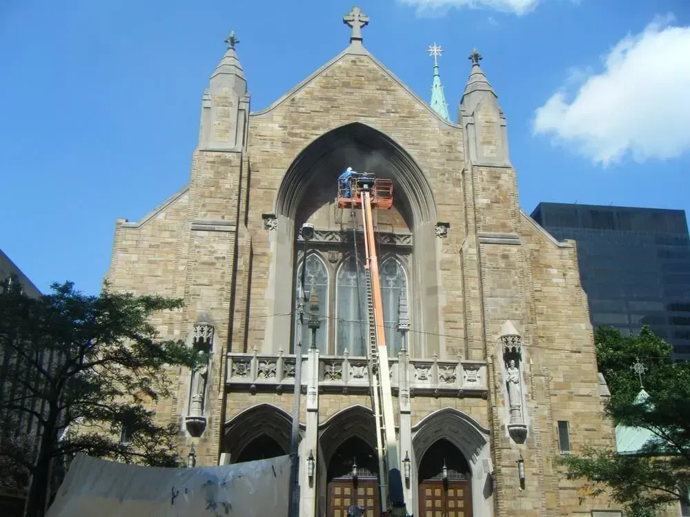 A worker in a lift repairs the facade of a stone church with a cross, against a blue sky.