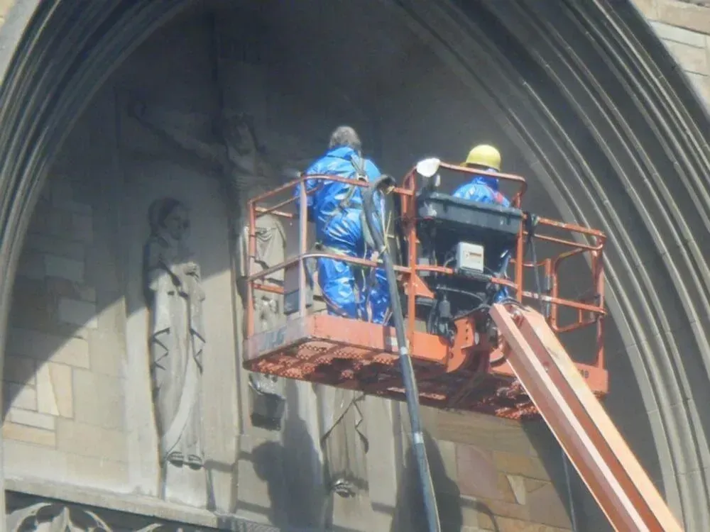 Two workers in protective gear on a lift, cleaning a church facade.