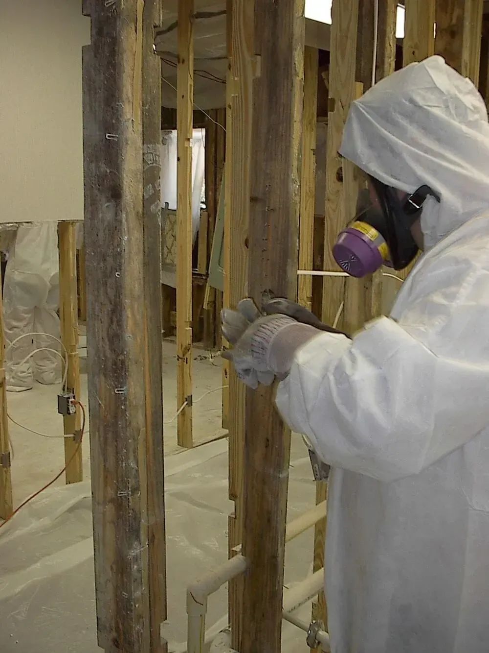Person in protective suit spraying wood frame. Indoors, white walls, and debris on the floor.