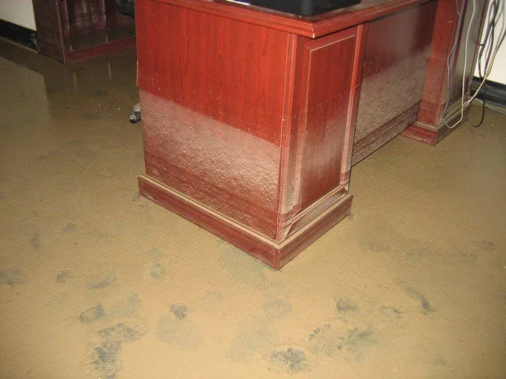 Floodwater surrounds a wooden desk in a room. Brown water stains the desk and floor.