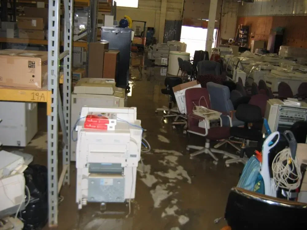 Warehouse flooded with water, various office equipment like printers and chairs, along with shelving units.
