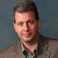 Man in a gray blazer and teal button-down shirt, looking directly at the camera with a neutral expression.