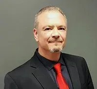 Man in black suit and red tie, smiling slightly against a gray background.
