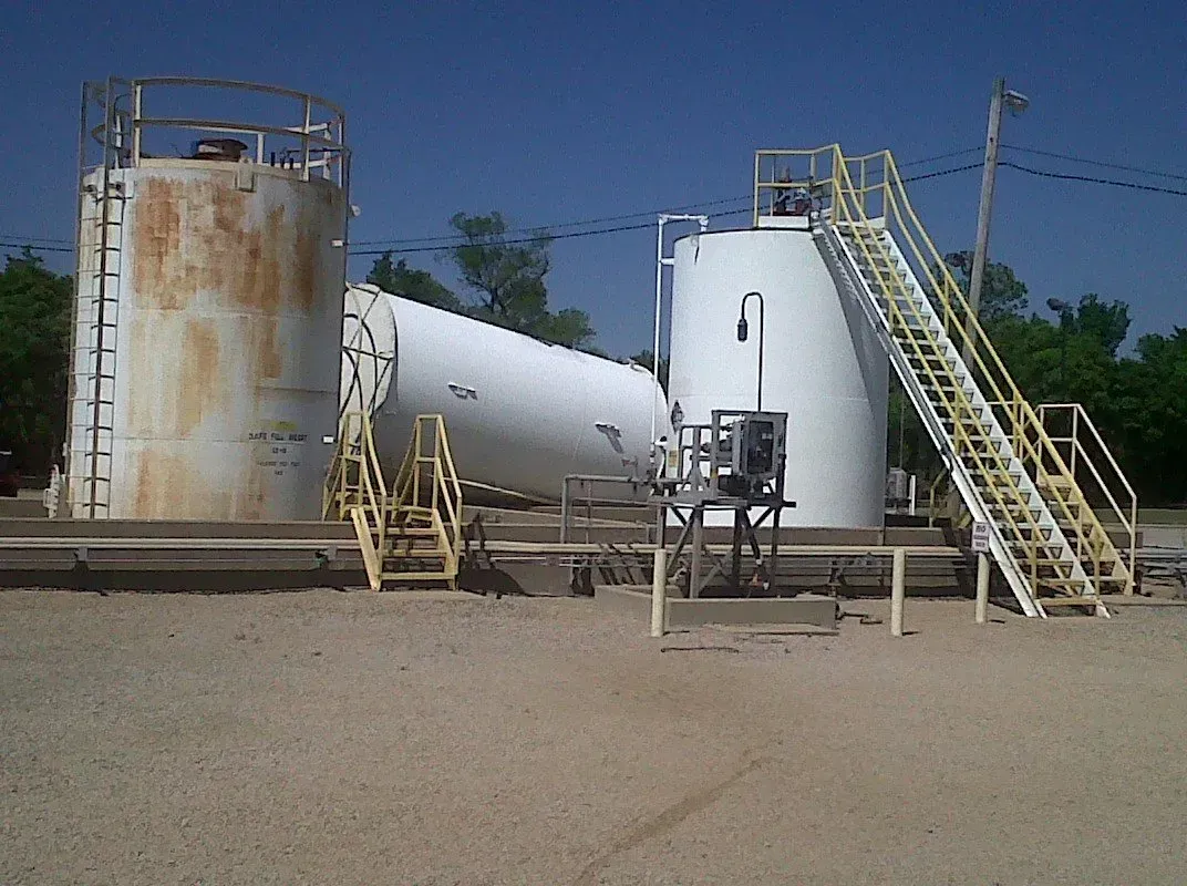 Oil storage tanks, two vertical, one horizontal, on a gravel surface under a blue sky.