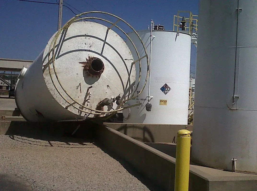 Overturned industrial tank; white with a circular access ladder, on a concrete pad with two other tanks.