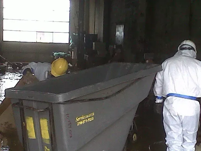 Workers in protective suits and a hard hat cleaning a debris-filled industrial area, large gray bin in the foreground.