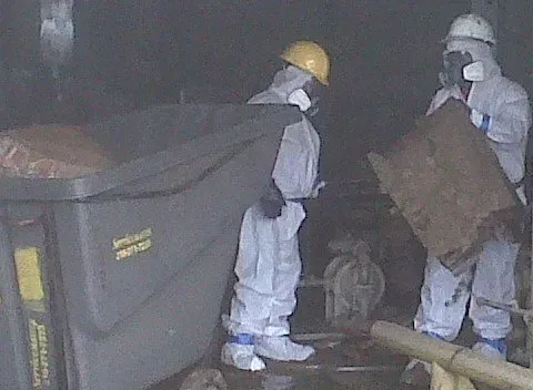 Two people in hazmat suits near a large trash bin and debris in a dim industrial setting.